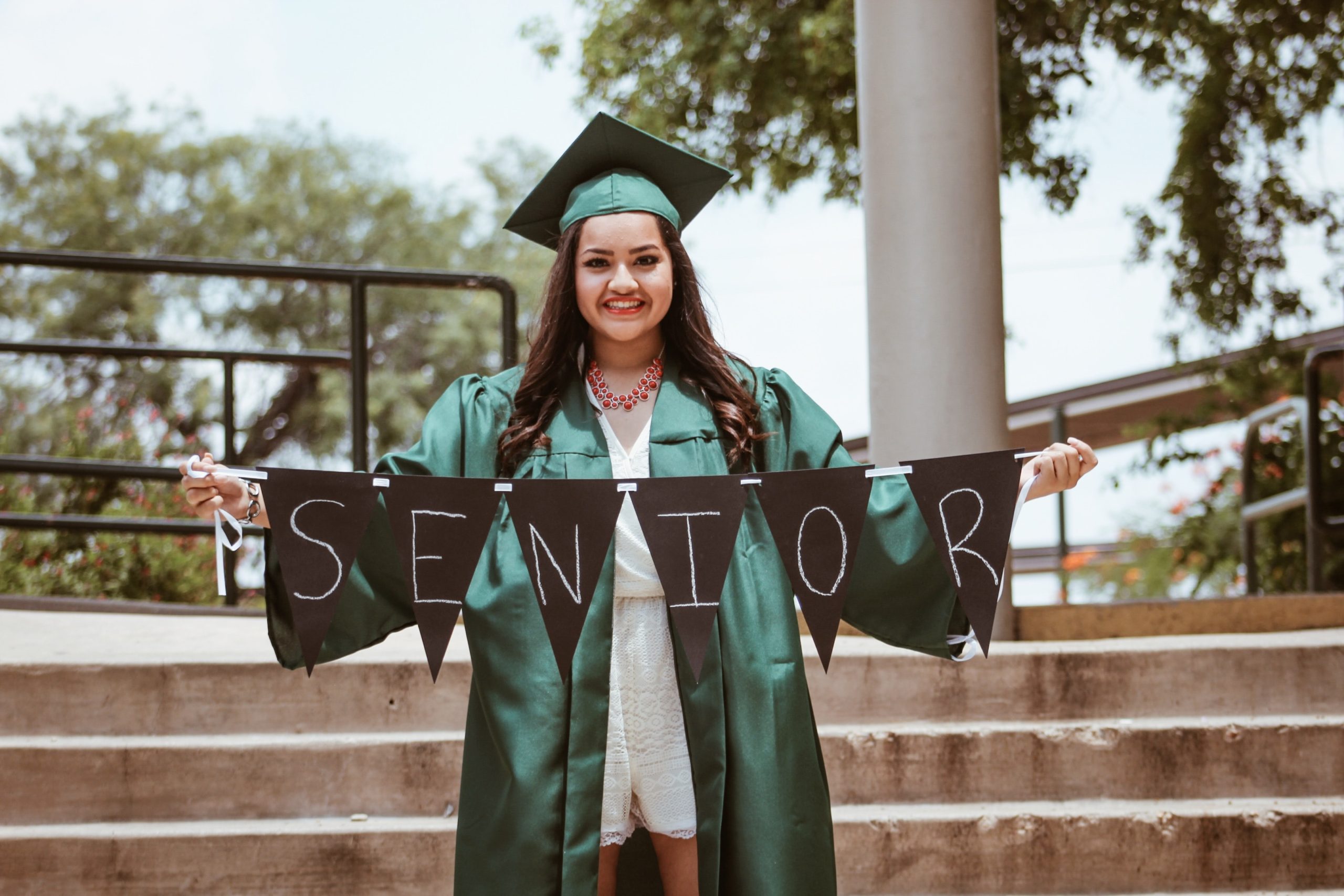 Girl in a green graduation gown holding up a sign that says seniors
